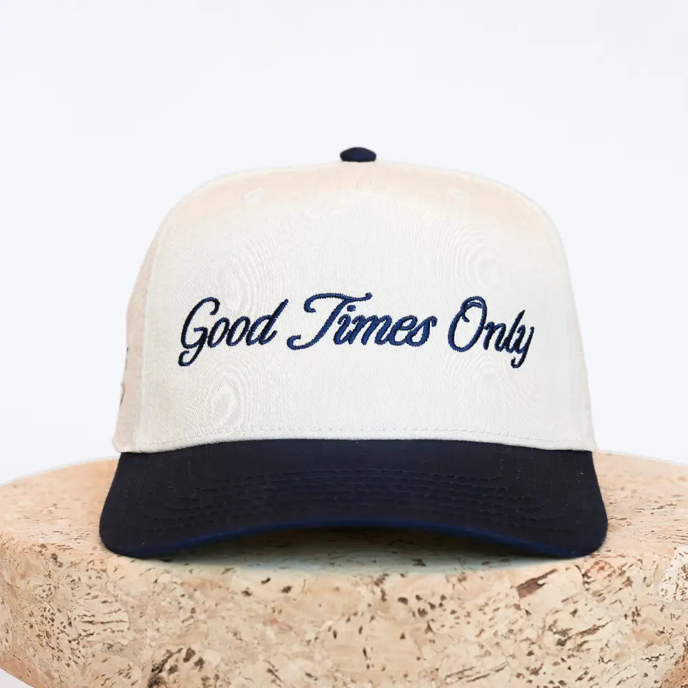 Cream-colored baseball cap with navy blue brim and embroidered ’good times only’ in cursive script.