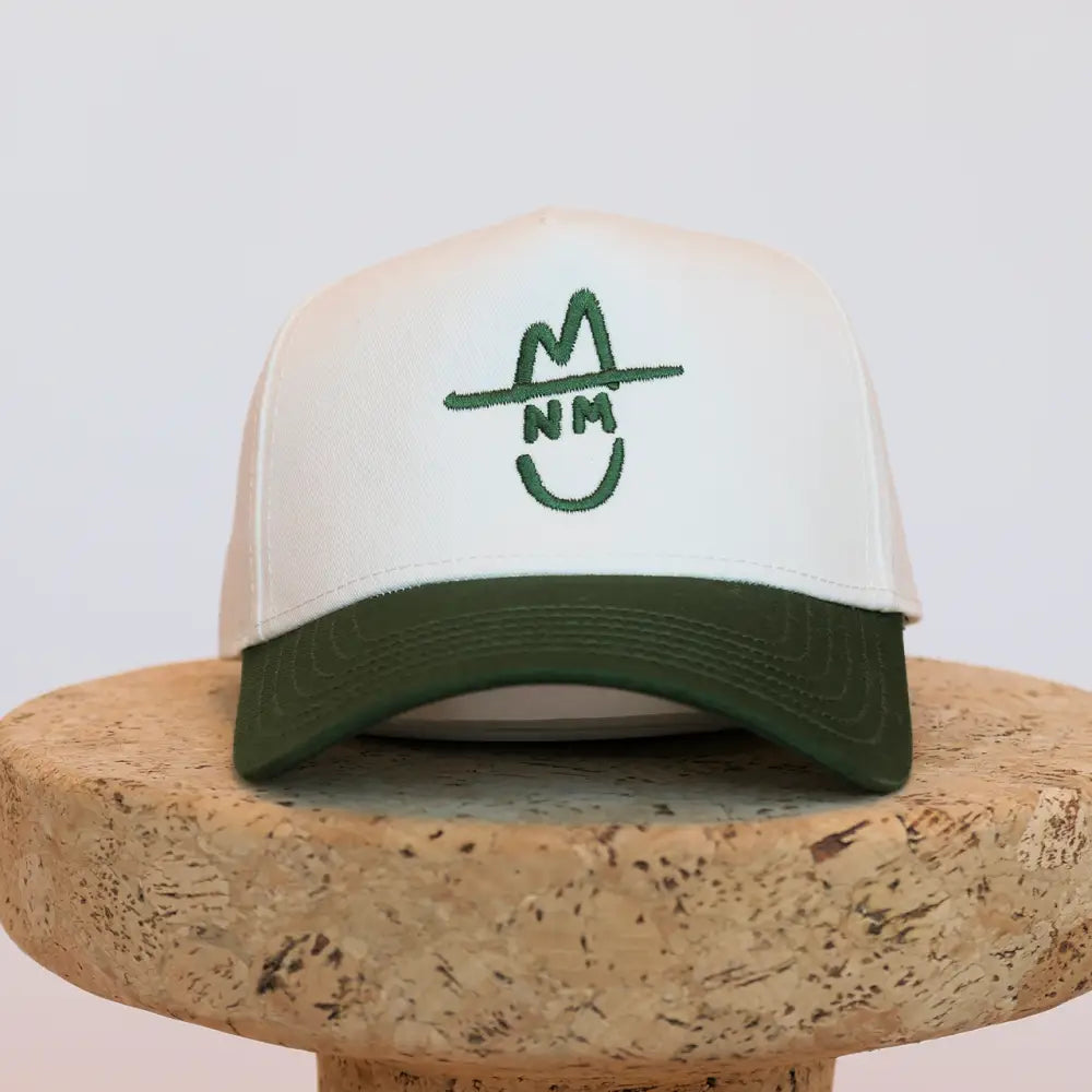 White and olive green trucker hat with a stitched logo of a smiling face wearing a hat.
